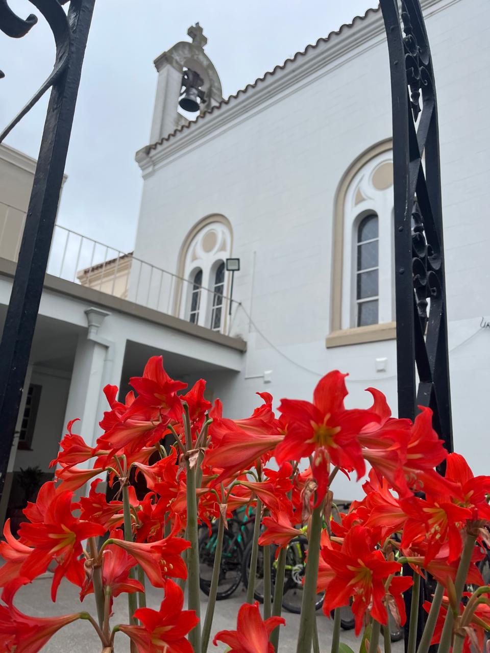 Capilla y campanario vistos desde la reja de entrada, con flores rojas